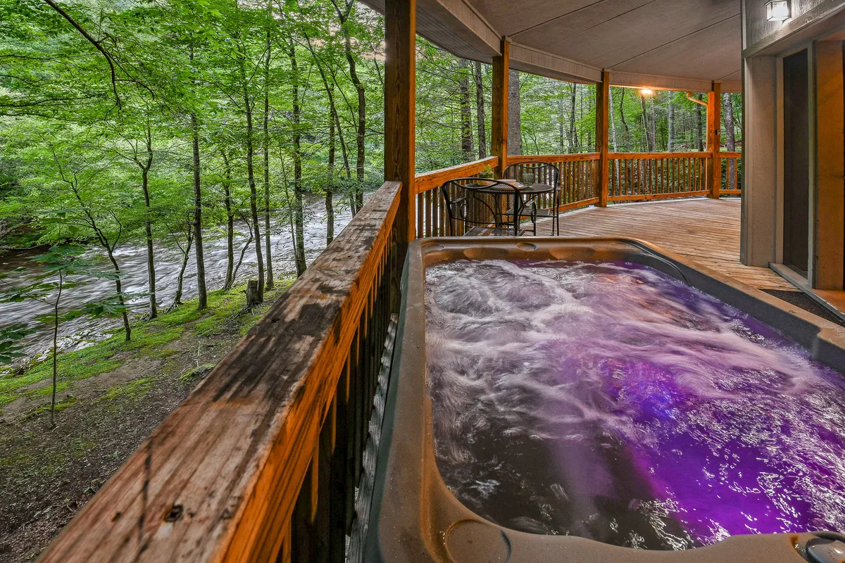 This serene exterior image features a private hot tub nestled on a wooden deck, surrounded by lush trees and overlooking the river. The tranquil setting exudes privacy, with the soothing sounds of water and a perfect spot for relaxing under the stars.