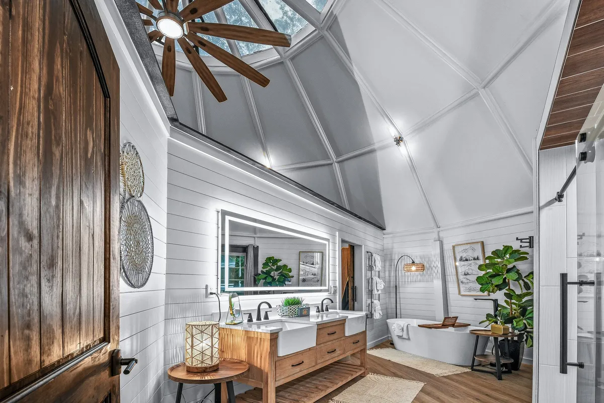 A serene, modern bathroom with a double vanity, expansive mirror, contemporary fixtures, a standalone bathtub, and touches of greenery and natural wood. Sunlight streams through a skylight, creating a bright and rejuvenating space designed for relaxation and self-care.
