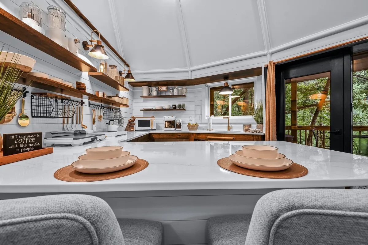 A warmly lit kitchen area showcasing a kitchen island set for breakfast, shelves with kitchenware, and large windows bringing in greenery and natural light. The setup hints at both meal preparation and leisurely dining, with a sense of openness and community.
