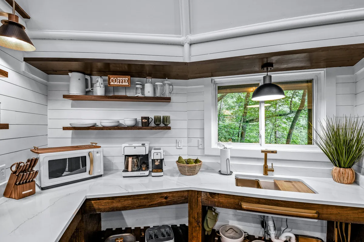 A close-up view of the kitchen counter featuring a coffee maker, wooden shelves lined with kitchen essentials, and a window framing a view of the surrounding forest. The setup creates a cozy, inviting space ideal for morning coffee rituals.