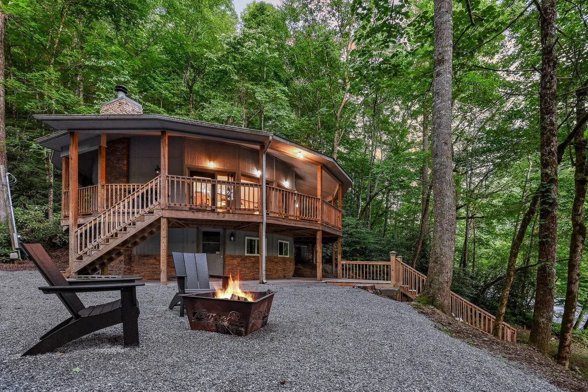 A circular wooden deck nestled among tall trees, featuring a central fire pit with outdoor seating arranged around it. Steps lead up to the deck and the surrounding natural landscape creates a peaceful, inviting atmosphere ideal for relaxation or social gatherings.