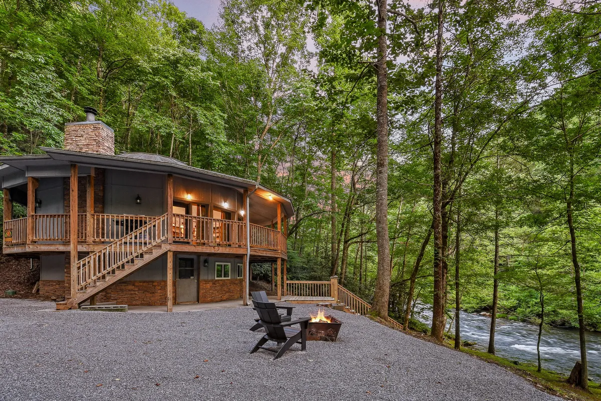 An inviting exterior scene showing the unique circular house, a cozy fire pit setup, and the surrounding trees with the river visible in the distance. The space is perfect for couples to gather around the fire under the night sky, enjoying the natural ambiance and connection to the outdoors.