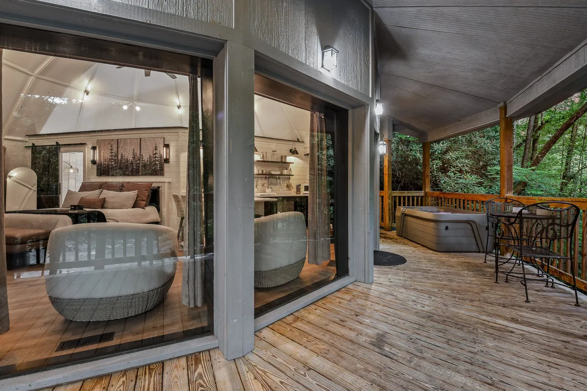 A cozy scene featuring large windows that reveal a comfortable interior, a porch with seating, and a prominent hot tub on the deck. The view emphasizes both the inviting indoor comforts and the easy access to outdoor relaxation.