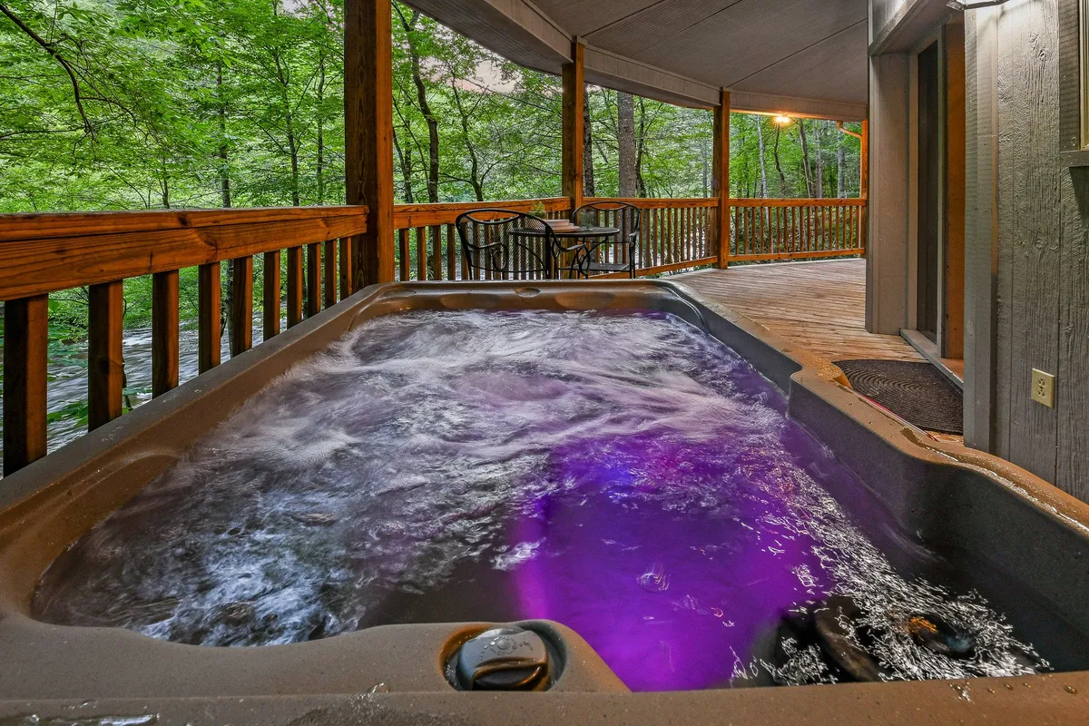An inviting outdoor hot tub with illuminated water sits on a wooden deck, surrounded by nature. The deck features a railing and an outdoor seating area, all set against the backdrop of tranquil greenery.