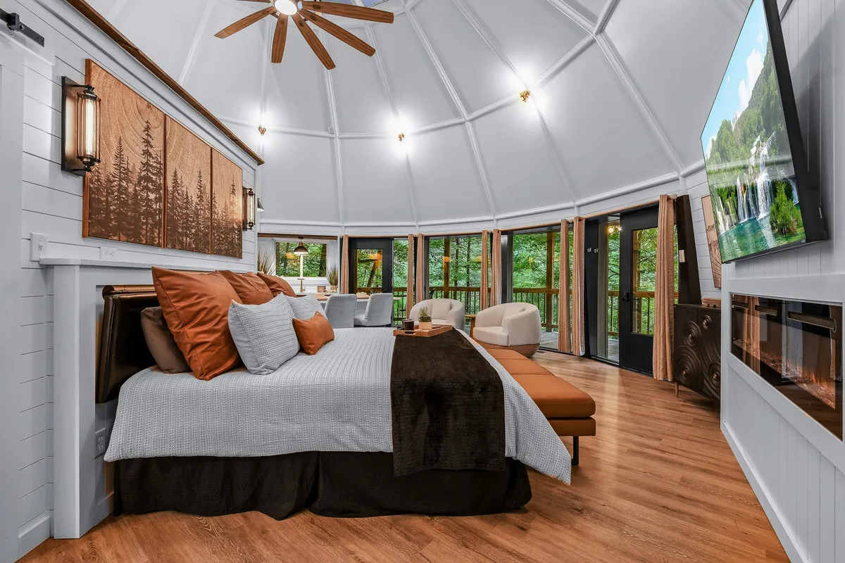 A bright, welcoming interior view with a bed adorned with decorative pillows, a TV mounted on the wall, a modern fireplace, and large windows that invite sunlight and nature views into the space. Warm wood accents and soft lighting create an intimate setting ideal for relaxation and connection.