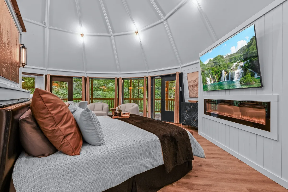 A tranquil studio view that captures the sleeping area with a cozy bed, large windows, a modern fireplace, and a flat-screen TV. The space feels airy and open, with the bed positioned to enjoy both the warmth of the fireplace and expansive mountain views through the windows. Soft, inviting lighting enhances the sense of closeness and comfort.