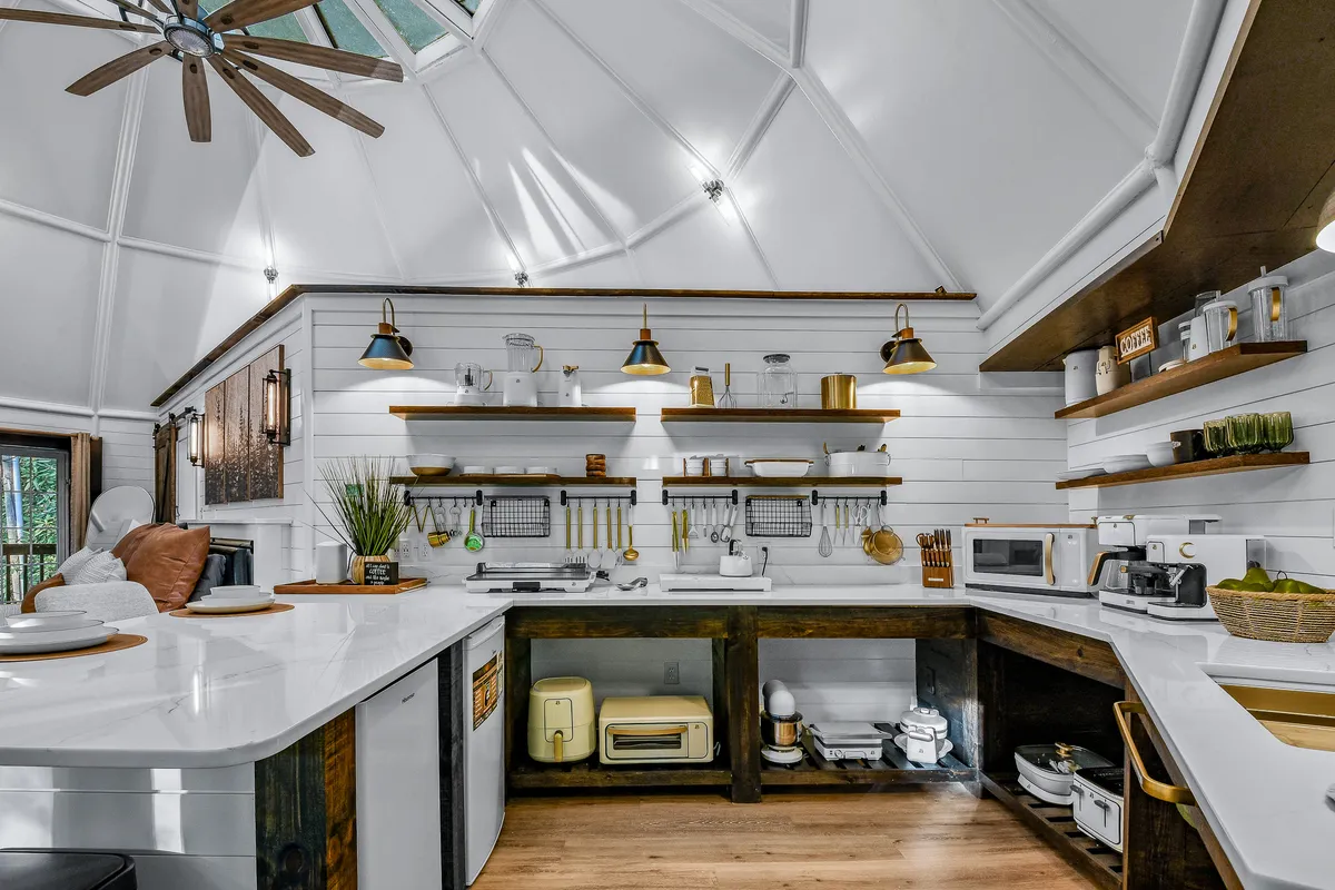 A welcoming kitchen scene centered on a large island with a pristine white countertop, accented by industrial-chic pendant lighting. Open shelving displays kitchenware, and wooden accents add warmth. The kitchen's open layout is visible, hinting at ample space for conversation and shared activities.