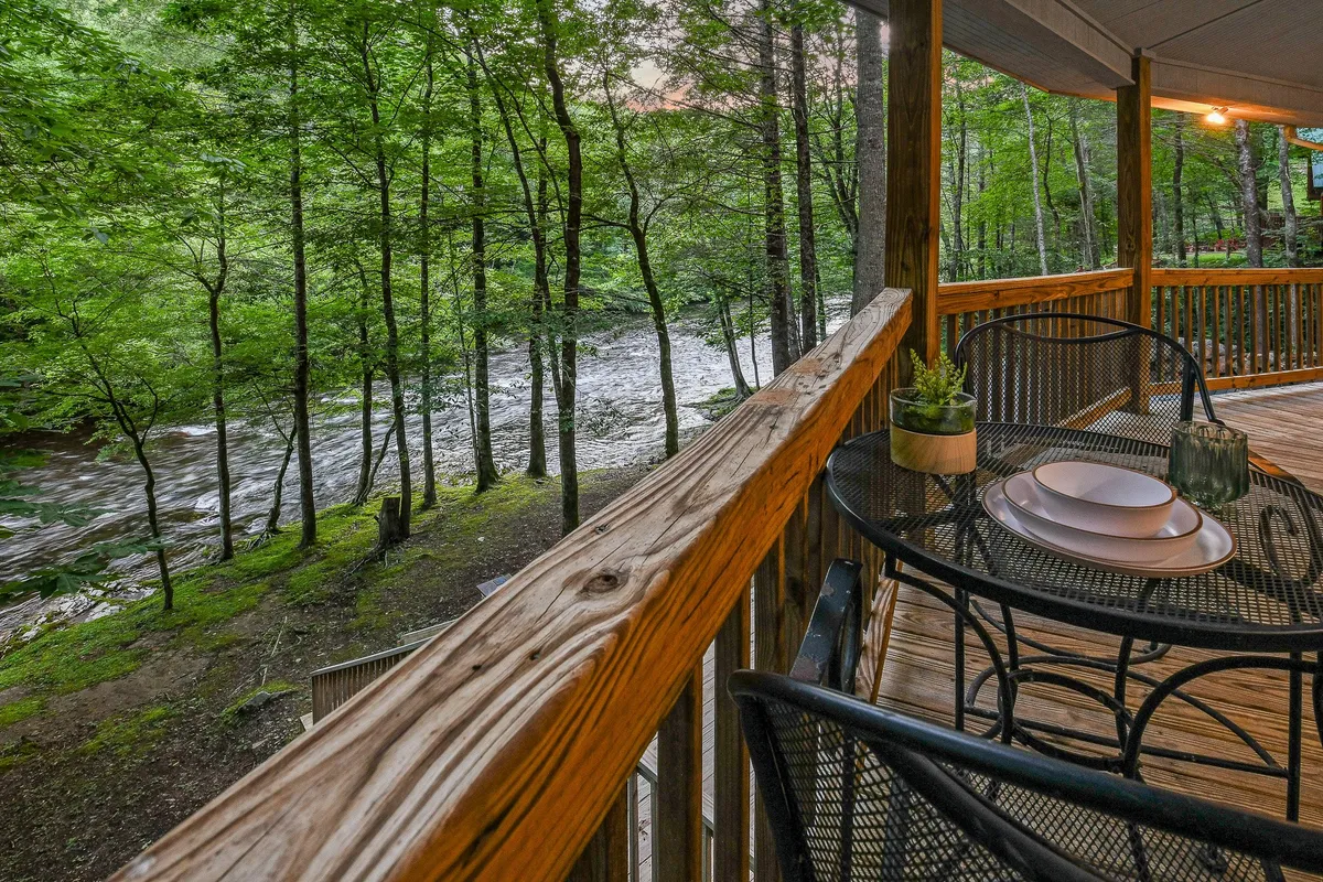 A peaceful riverside deck with a wooden railing, outdoor dining set, and sweeping views of the flowing river and trees. The scene is ideal for morning coffee, outdoor meals, and quietly taking in the natural landscape just steps from the cottage.