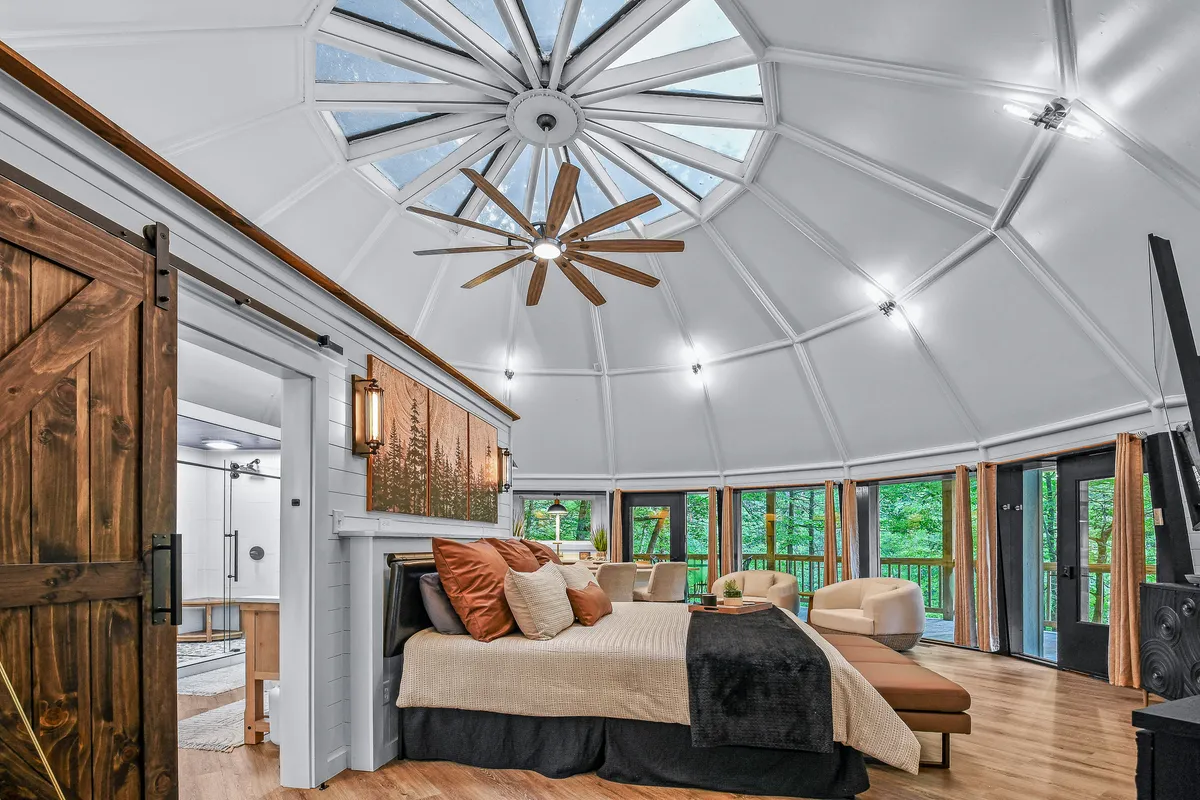 A spacious interior featuring a central king-size bed with decorative pillows, set beneath a striking circular domed ceiling with skylights. Large panoramic windows bathe the space in natural light, while a cozy seating area and rustic wooden barn door complete the look. The ambiance is calm and inviting, with clear sightlines toward the surrounding nature.