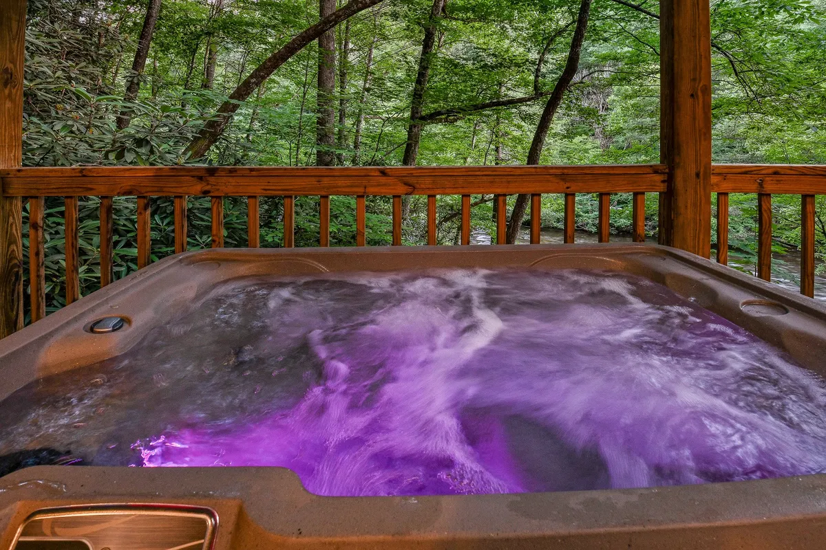 This exterior image features a hot tub with vibrant purple lighting set on a wooden deck, surrounded by trees and a rustic wooden railing. The natural backdrop and ambient lighting create a tranquil atmosphere, inviting guests to unwind while staying connected to the outdoors.